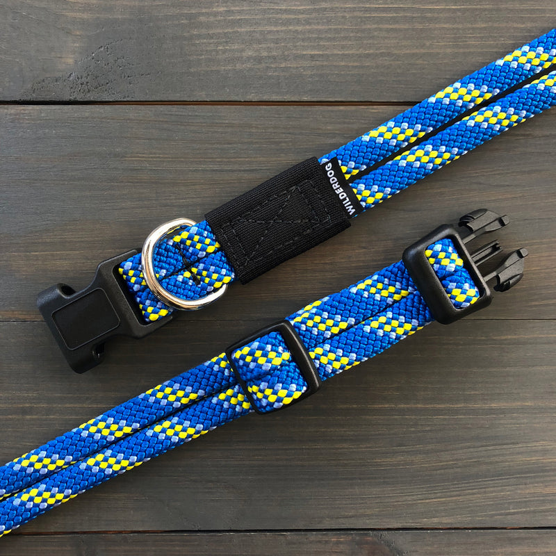 The Mariner Collar set, featuring a vibrant dog leash and matching collar, is displayed on a dark wooden surface. The leash is crafted from blue, yellow, and white woven fabric and includes black plastic buckles and a metal ring attachment. A black fabric tag with white text is securely attached to the durable leash near the buckle.